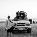 Black and white photo of a 4x4 vehicle on a beach carrying surfboards, capturing a relaxed seaside vibe.